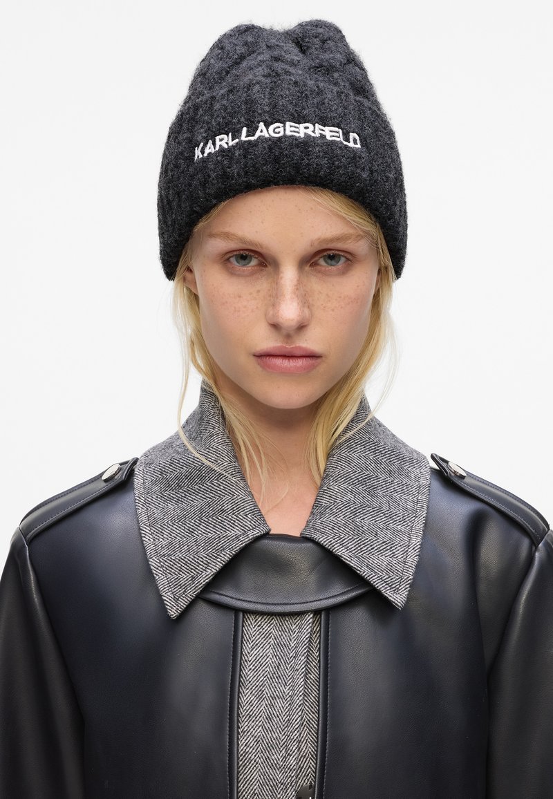 Gorro de lana de carbón con el logo "KARL LAGERFELD", combinado con una chaqueta de cuero negra que presenta un cuello de espiga gris y un tejido texturizado.