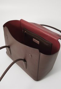 Borsa tote in pelle marrone con una finitura texturizzata, caratterizzata da un interno rosso cupo e una tasca con zip. Manici morbidi e arrotondati si estendono dai lati.