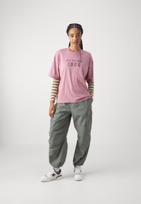 Roza bombažna majica s printom "New Balance 1906", kombinirana z zelenimi cargo hlačami. Model nosi bele in vijolične superge. Vidne so črtaste rokave.