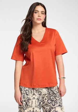 Frau mit langen dunklen Haaren trägt ein lockeres orangefarbenes Kurzarmshirt mit V-Ausschnitt und cremefarbene Hose mit Blumenmuster vor einem unifarbenen Hintergrund.