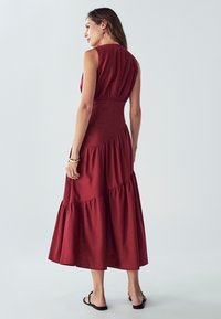 Rdeča brezrokavna midi obleka s slojevitim robom, nabrano pasom in gumbom na hrbtni strani. Mehka tkanina z gladko teksturo.
