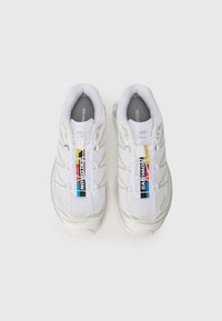 Zapatillas deportivas blancas con parte superior de malla, suelas de goma y etiquetas de marca coloridas. Presentan un detalle texturizado y un sistema de cordones en la parte delantera.