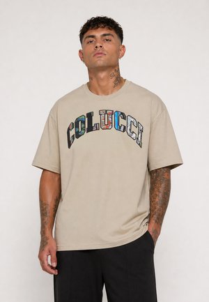 Man met tatoeages draagt een beige oversized T-shirt met kleurrijke "COLUCCI" tekst en zwarte broek, staand tegen een effen lichte achtergrond.