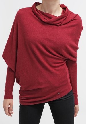 Persona con un suéter rojo de punto de manga larga y holgado, con un escote ancho y caído y mangas ajustadas, combinado con pantalones negros.