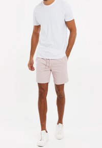 Homme debout portant un t-shirt blanc simple, un short rose clair avec cordon de serrage et des baskets blanches sur un fond blanc.