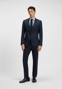 Marineblauw kostuum met een slim fit, voorzien van een one-button blazer, met revers en een gestreept overhemd eronder. Zwarte schoenen.