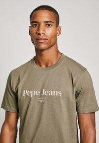 Olivově zelené bavlněné tričko s krátkým rukávem, na němž se nachází krémové logo "Pepe Jeans", kulatý výstřih a hladká textura.