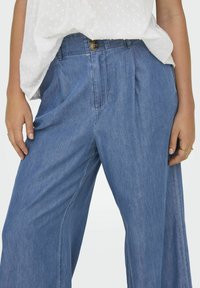 Pantalones de mezclilla anchos de color azul claro con pliegues, que presentan un cierre de botón en bronce y una textura sutil. Se llevan con una blusa blanca.