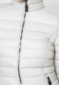 Jasnoszara kurtka puchowa z wysokim kołnierzem, poziomym przeszyciem i przednim zamkiem błyskawicznym z kontrastującym czarnym uchwytem.