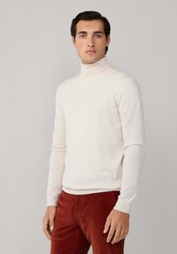 Cremfarbener Rollkragenpullover aus weichem Strickmaterial, kombiniert mit rostroten Cordhosen, die eine taillierte Passform und gerippte Bündchen haben.