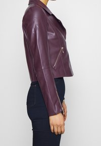 Veste de motard en cuir bordeaux avec un design court, présentant un col cranté, des poches zippées et une finition brillante.