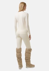 Vrouw in een strakke crèmekleurige lange mouw top en legging met beige gewatteerde winterlaarzen, met de rug naar de camera gekeerd tegen een lichte achtergrond.