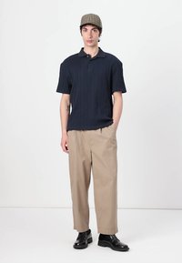 Navy ribbed short-sleeve polo shirt, beige wide-leg trousers, and black leather shoes. Model wears a plaid cap. Minimalist backdrop.