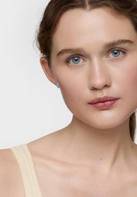 Primo piano di una persona con pelle chiara e occhi blu. Indossa piccoli orecchini blu; i capelli sono castani e leggermente ondulati, con uno sfondo neutro.