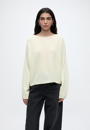 Frau mit langen dunklen Haaren, die goldene Creolen, einen locker sitzenden cremefarbenen Pullover und schwarze weite Hosen trägt und vor einem hellen Hintergrund steht.