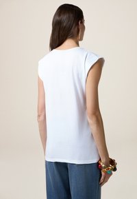 T-shirt senza maniche in cotone bianco con scollatura rotonda, texture liscia e vestibilità comoda, abbinata a jeans e bracciali colorati.