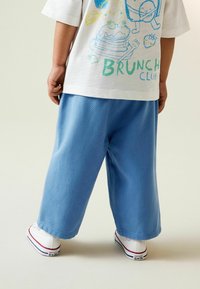 Helehallid laia lõikega püksid, mis on valmistatud pehmest kangast, koos valge T-särgiga, millel on värvikad brunch-teemalised graafikad. Valged tossud punaste aksentidega.