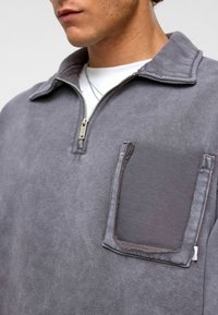 Grijze pullover met een ritssluiting bij de kraag, gemaakt van een gestructureerde stof, met een patchzak op de borst en zilverkleurige hardware op de rits.