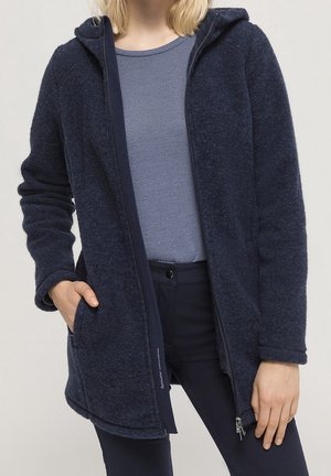 Femme portant une veste polaire bleu foncé zippée avec capuche, superposée sur un haut bleu clair texturé et un pantalon foncé, main dans la poche.