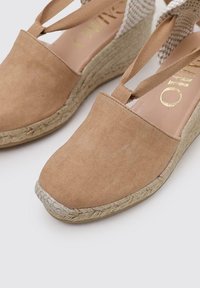 Espadrilles de cuña de lona en tono beige con un tacón de acento trenzado y diseño de tiras cruzadas. Los detalles de cuerda a lo largo de la suela añaden textura.