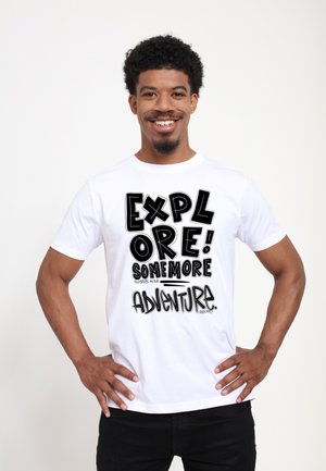 Lachende man met krullend haar, die een wit T-shirt draagt met de tekst "Explore! Some more adventure", staand met handen in de zij.