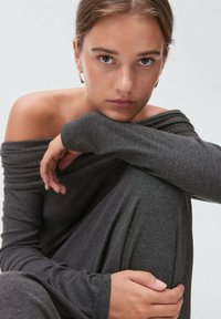 Top s odhalenými ramenami a dlhými rukávmi v tmavošedej pletenej látke. Mäkky textúr a voľný strih s ryhačnými detailmi na rukávoch.