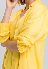 Chemise jaune à manches retroussées, en tissu léger, avec une texture lisse et un débardeur assorti en dessous.