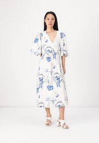 Weißes Wickelkleid mit blauen Blumen- und geometrischen Mustern; verfügt über kurze, gepuffte Ärmel und eine betonte Taille. Kombiniert mit weißen Blockabsatzsandalen.