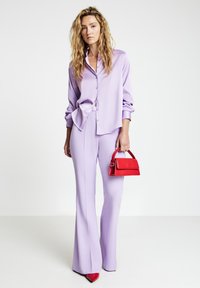 Woman with long curly hair wearing a lavender button-up shirt and pants, holding a small red handbag and wearing red pointed shoes.