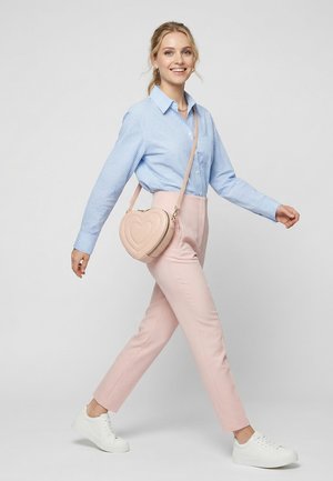 Woman wearing light blue checkered shirt and pink trousers, walking with a heart-shaped beige crossbody bag and white sneakers.