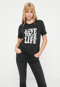 Μαύρο βαμβακερό t-shirt με λευκή εκτύπωση που γράφει «LOVE YOUR LIFE» με έντονη γραμματοσειρά, σε συνδυασμό με σκούρο τζιν παντελόνι, τα χέρια στις τσέπες.
