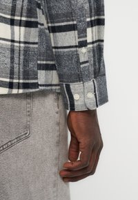 Αριστερό χέρι σε μακρυμάνικο πουκάμισο με καρό μοτίβο σε γκρι, μαύρο και λευκό χρώμα, με τη μανσέτα πάνω στην τσέπη ενός ανοιχτό γκρι τζιν παντελονιού σε κοντινό πλάνο.