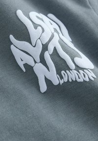 Sivá látka s vystupujúcim bielym textom „ASAYAN LONDON“ v tučných, nepravidelných písmenách s textúrovaným, trojrozmerným efektom.
