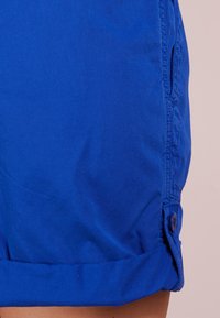 Ljusa blå shorts med upprullade fållar och sidoknappar på en ljus bakgrund, som visar en del av ett ben.