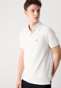 Jonge man met krullend haar die een wit poloshirt met korte mouwen draagt met een klein rood "cK"-logo op de borst, kijkt opzij.