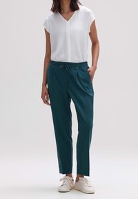 Pantalones verdes con cintura elástica y piernas ajustadas, combinados con una blusa blanca de manga corta, usados con zapatillas blancas. Tela suave con un ajuste relajado.
