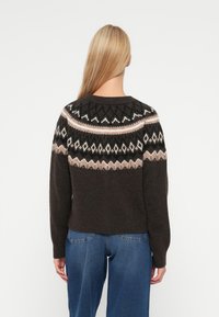 Pulover tricotat de culoare maro închis, cu un model geometric pe umerii din crem și roz pal. Are un gât rotund și umeri lăsați.