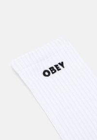 Baltos šonkačiuotos kojinės su juodu siuvinėjimu „OBEY“ ant krašto. Audinys atrodo minkštas su tekstūruotu paviršiumi.