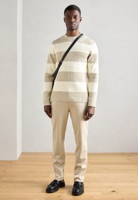 Beige ja kerma triibuline ribav soe pusa pikade varrukatega, kombineeritud beežide pükstega ja mustade kingadega, kaasas must risti kott.