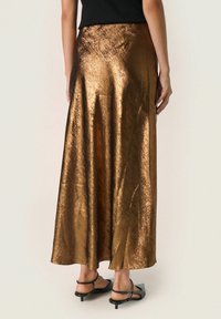 Maxi gonna in bronzo metallico con texture liscia e leggero luccichio, caratterizzata da una vestibilità comoda e orlo fluido. Indossata con scarpe nere.