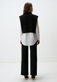 Zwarte gebreide vest met hoge kraag, gecombineerd met een lange witte blouse en wijde zwarte broek, gedragen met zwarte hakken. Eenvoudig en modern ontwerp.