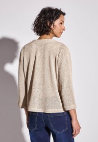 Beige stickad tröja med trekvartslånga ärmar, öppen ryggdetalj, texturerad tyg och en avslappnad passform, ihop med blå jeans.