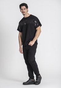 Camiseta negra de manga corta con un logotipo negro audaz, combinada con jeans oscuros y zapatillas negras texturizadas, creando un look monocromático.