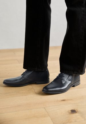 Zwarte dresschoenen met een gestructureerd oppervlak, puntige neus en vetersluiting, gecombineerd met zwarte jeans, geplaatst op een houten vloer.