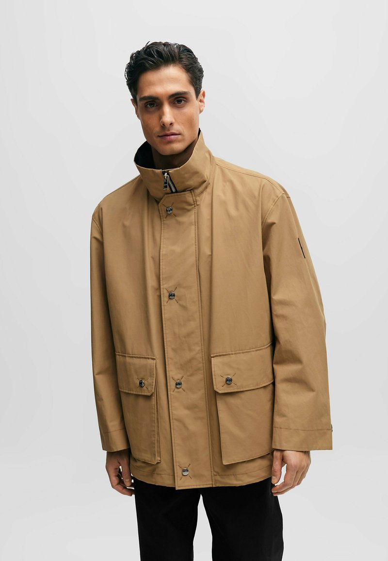 Mann mit dunklen Haaren, der eine sandfarbene Jacke mit hohem Kragen, Frontknöpfen und Klappentaschen trägt, und vor einem einfarbigen hellen Hintergrund steht.