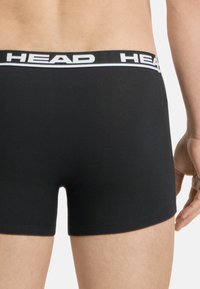 Boxeurs noirs avec une bande blanche affichant "HEAD", présentant une texture lisse et une coupe ajustée. La taille est élastique et contraste avec le tissu.