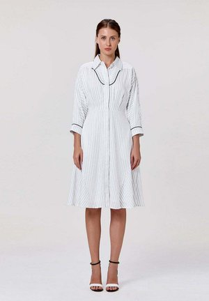 Femme debout face à nous portant une robe blanche au genou avec des rayures verticales et des bordures noires au col et aux poignets, associée à des talons blancs.