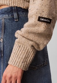 Μανίκι μπεζ πλεκτού πουλόβερ με ετικέτα Calvin Klein, φορεμένο μέσα σε ψηλόμεσιο μπλε τζιν παντελόνι.
