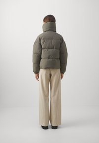 Person, der von hinten mit einer hochkragen olivgrünen Steppjacke und weiten beigen Cordhosen vor einem schlichten weißen Hintergrund steht.