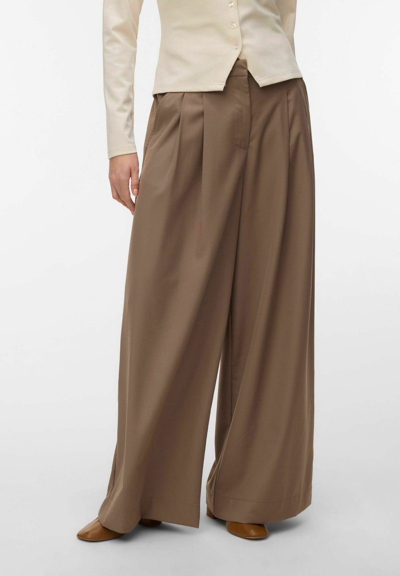 Femme portant un pantalon marron taille haute à jambes larges, associé à un haut crème à manches longues boutonné et des chaussures marron, debout devant un fond blanc.